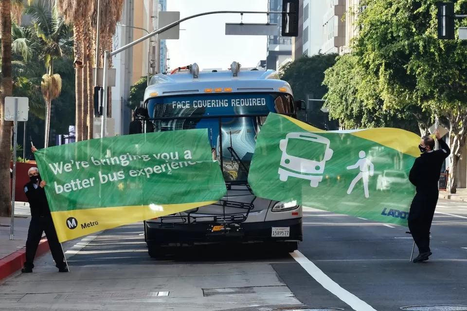 Bus driving through green banner held by two Metro employees at inauguration of new bus lane in downtown Los Angeles.